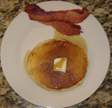 Finished pancakes on a plate with butter, maple syrup, and bacon.